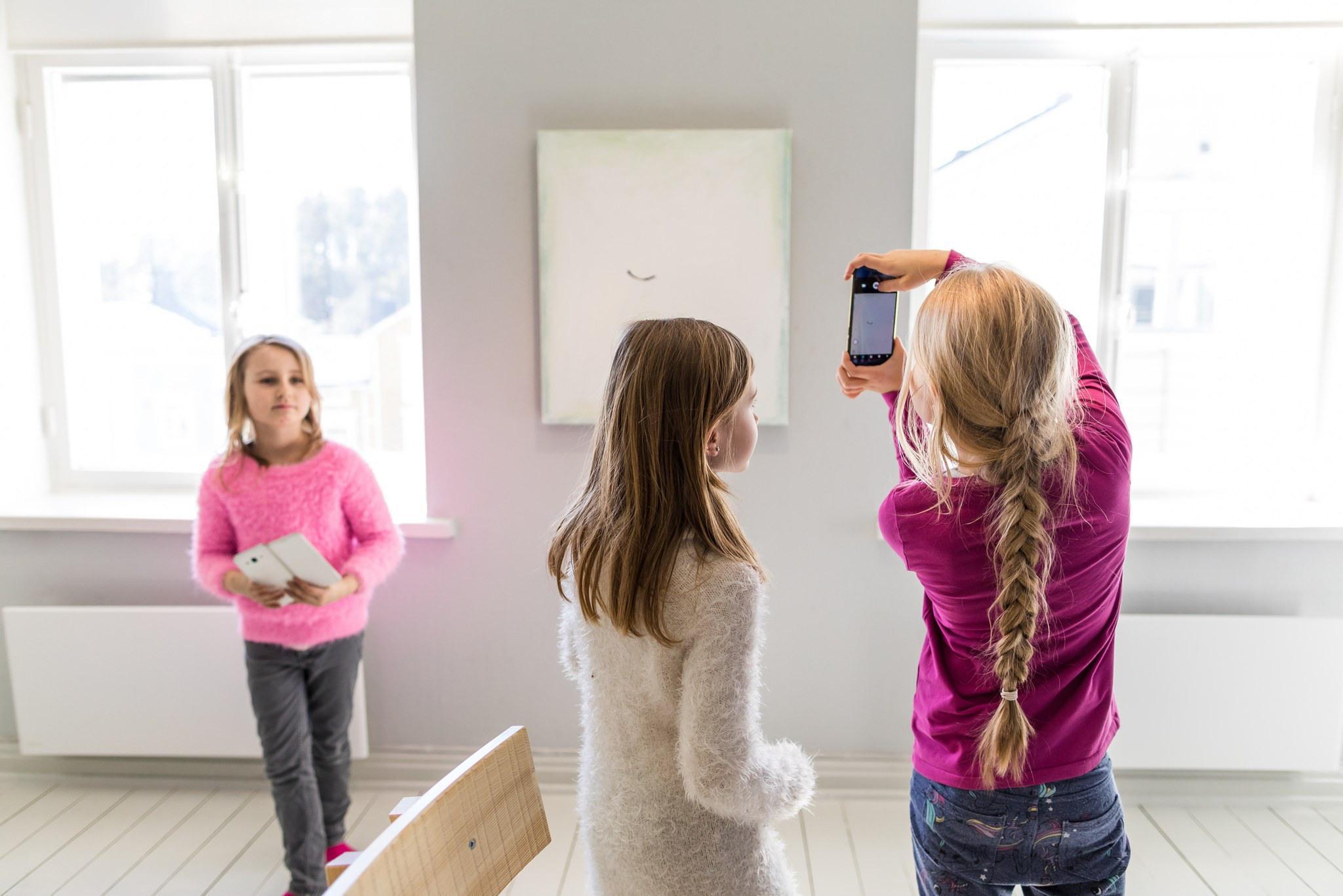 Kolmea alakouluikäistä tyttöä ottaa puhelimella kuvia teoksista taidenäyttelyssä