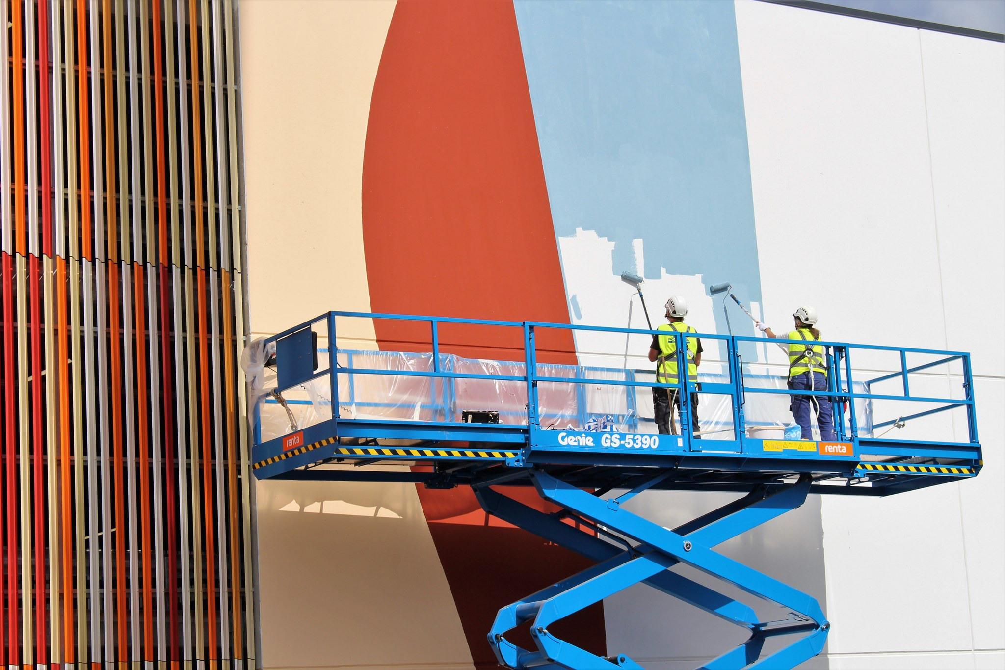 Kaksi henkilöä maalaavat valtavaa ja värikästä taideteosta rakennuksen ulkoseinää.