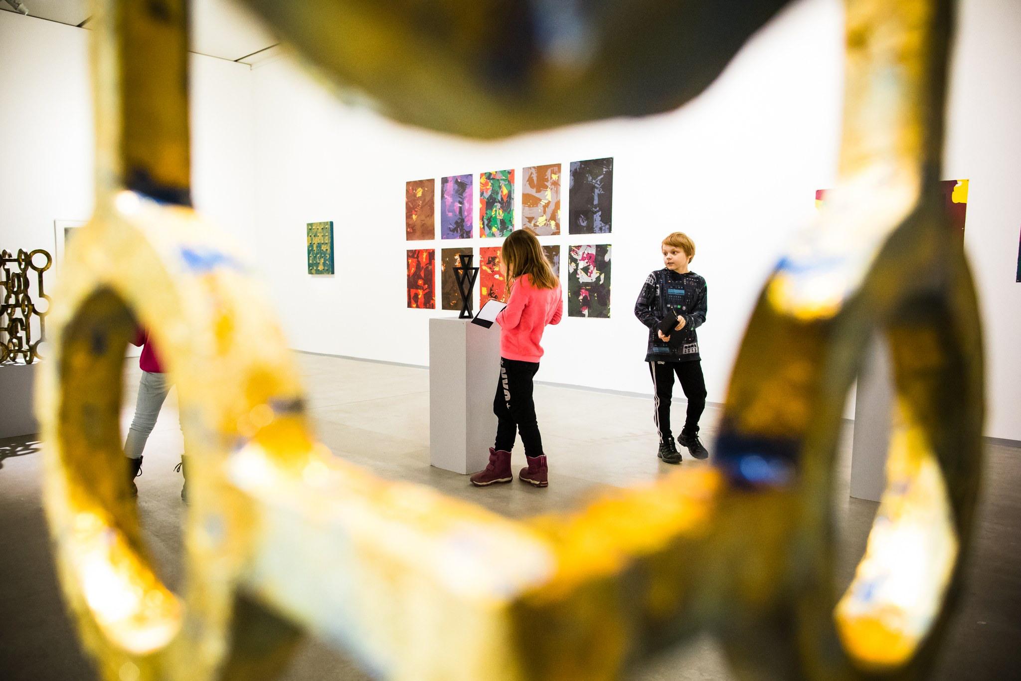 2 alakouluikäistä lasta tekevät muistiinpanoja värikkäiden taideteosten ympäröimänä isossa näyttelytilassa