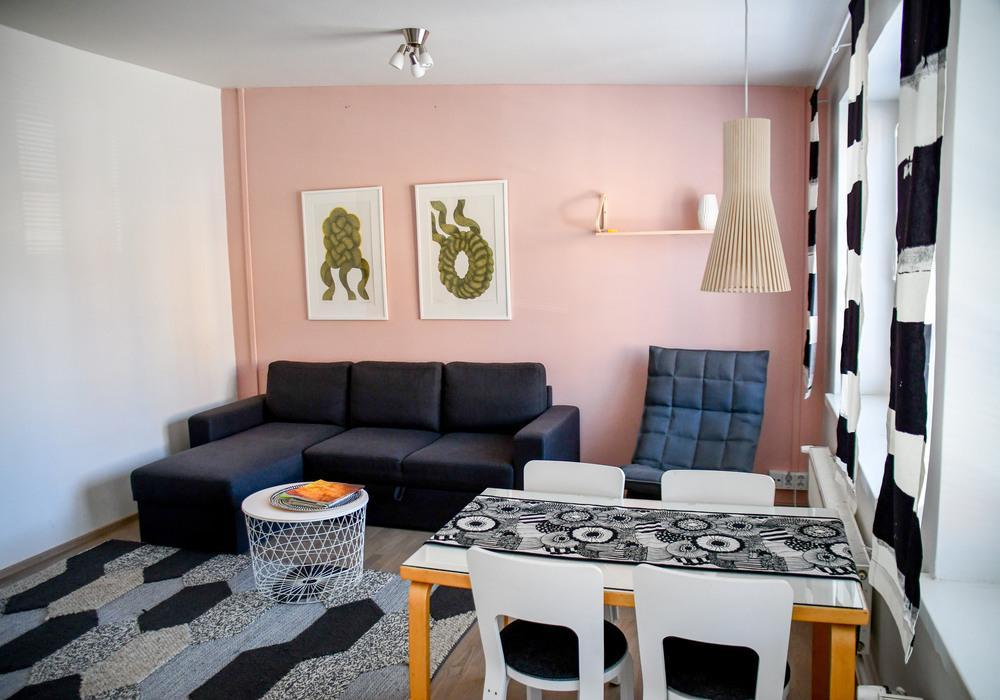 Kuva huoneesta jossa vaaleanpunainen takaseinä. Huone on sisustettu sohvalla, pöytäryhmällä ja nojatuolilla. Ikkunoissa roikkuu verhot ja seinällä kaksi taulua.