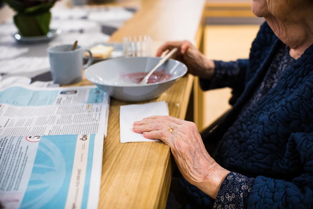 Äldre kvinna äter och läser dagstidning vid matbordet.