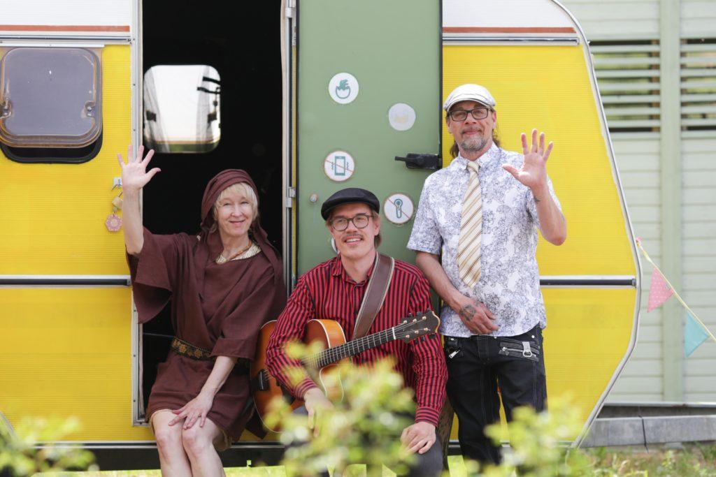 Tre artister sitter framför ett gult husvagn.