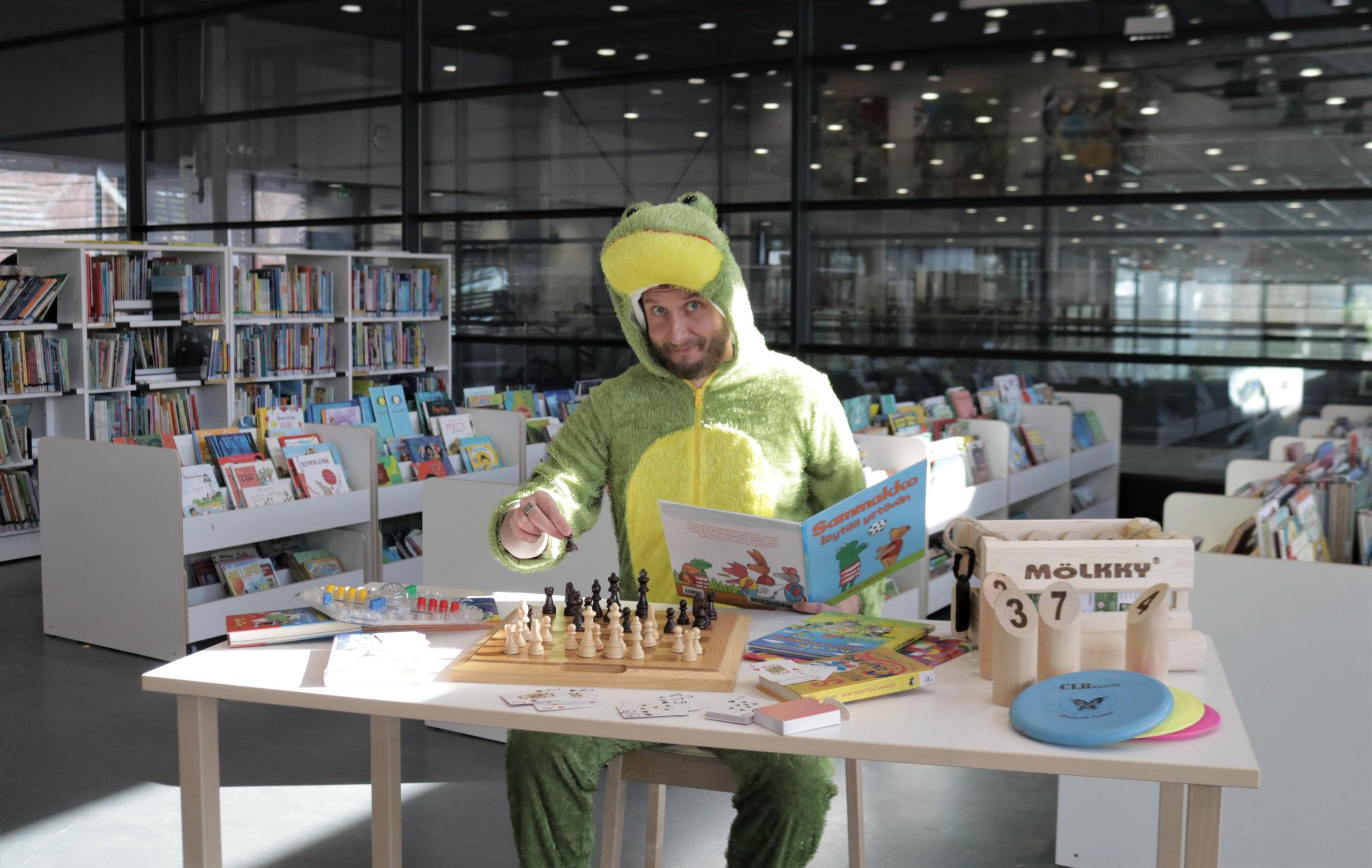Kirjastosammakko pelaa shakkia ja lukee kirjaa.