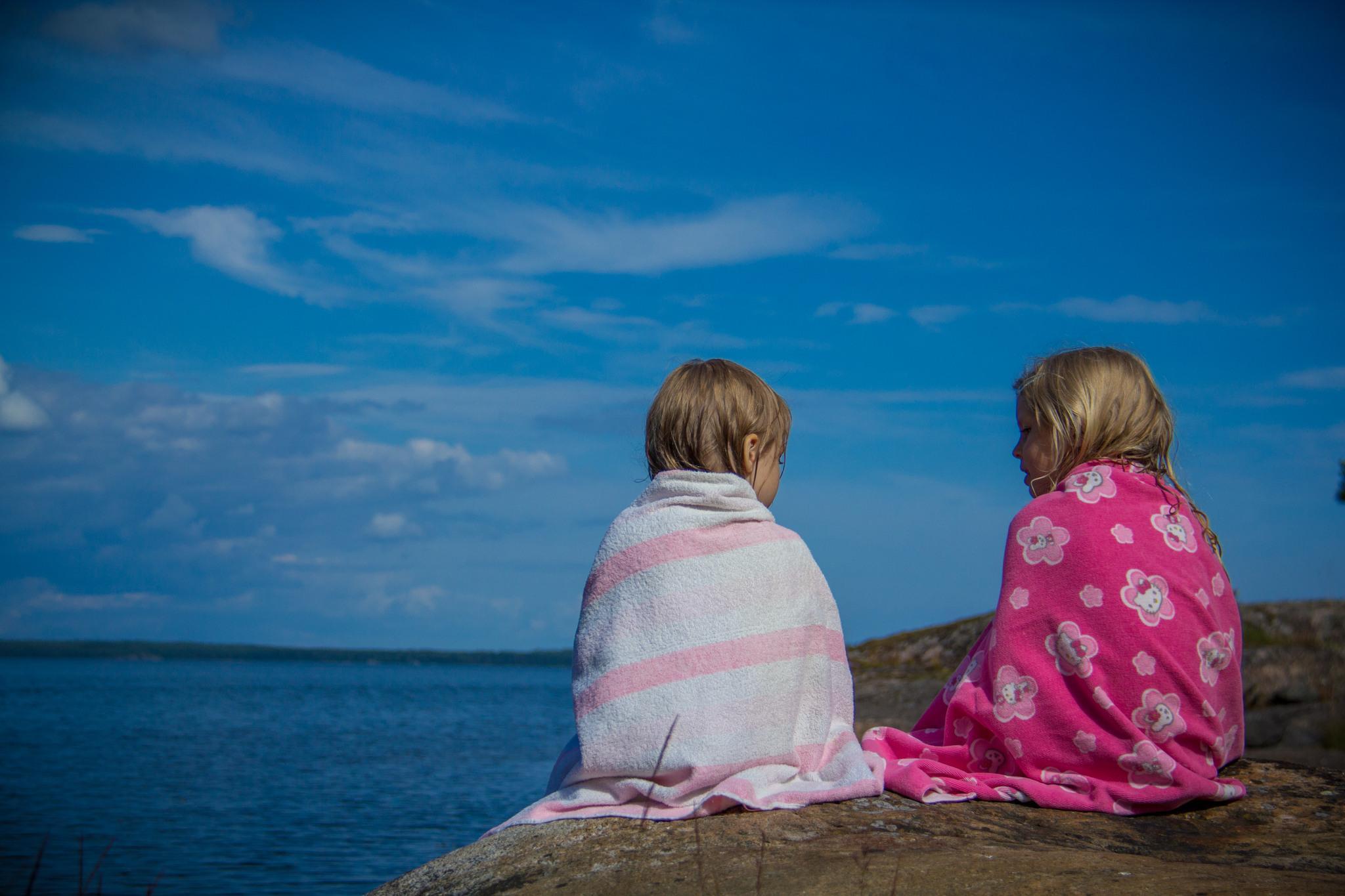 två barn med handdukar kring sig sitter på en klippa vid vattnet.