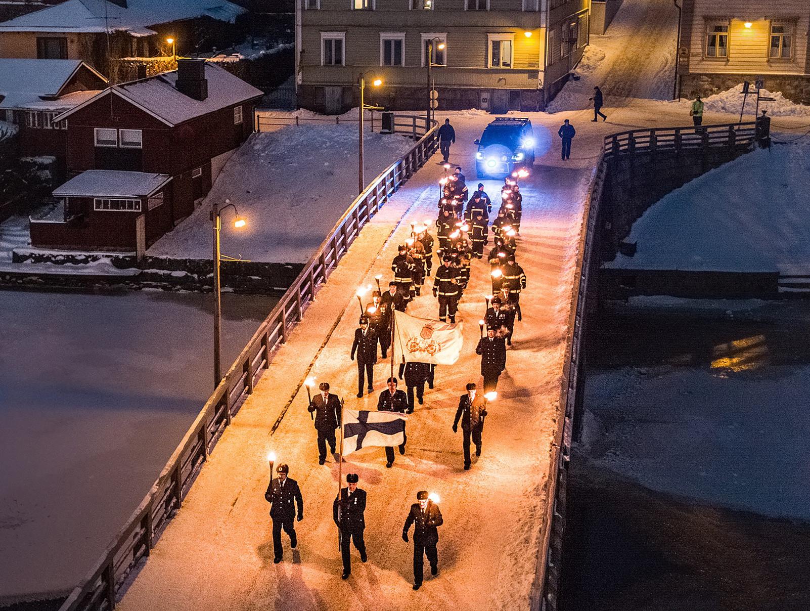 Runeberginpäivän soithukulkue kävelee Porvoon vanhan kaupungin sillan yli. Edessä mies joka pitää käsissään Suomen lippua.