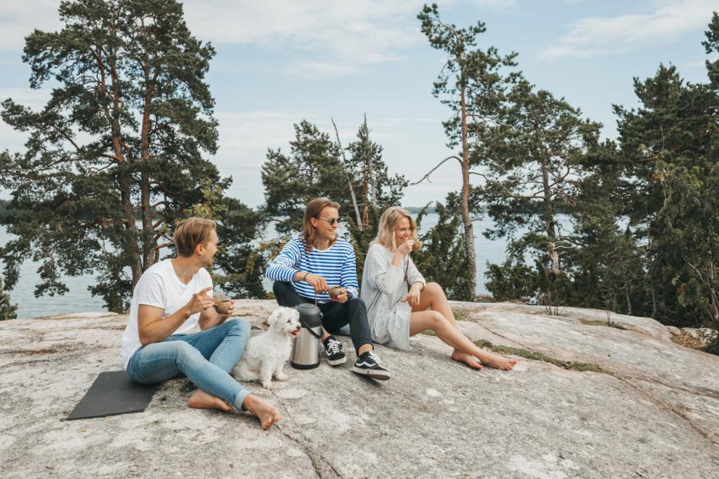 Kaksi miestä, nainen ja koira istuvat kalliolla saaristossa.