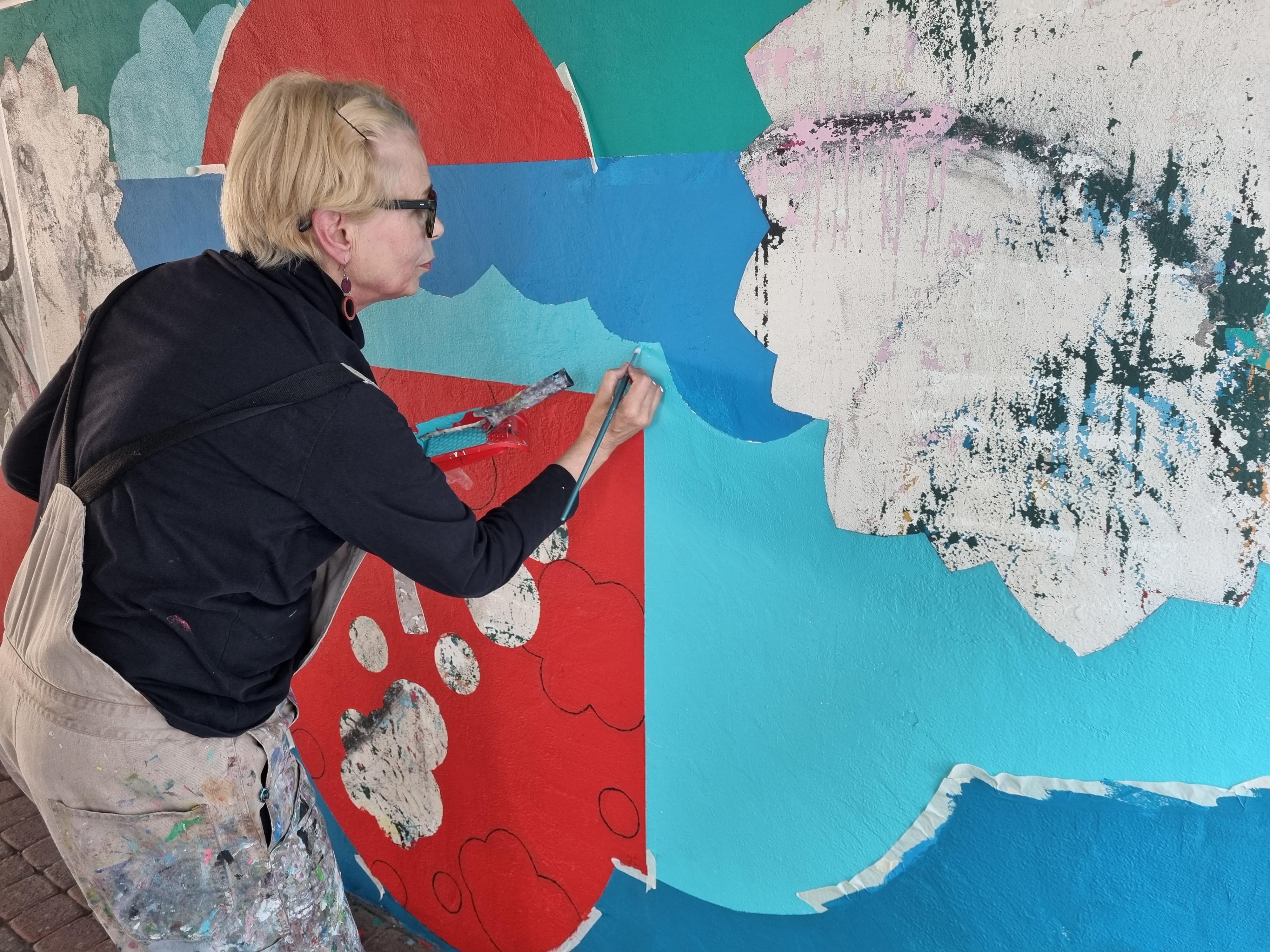 Konstnären Irmeli Mäkilä arbetar på en mural, hon målar med blå färg på en vägg.