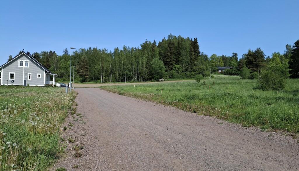 Maisema Carlanderintieltä: rakentamattomia tontteja tien molemmin puolin vanhalla peltoaukealla, vasemmalla yksi valmis omakotitalo, taustalla metsää. Valokuva.