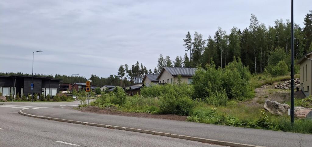Valokuva rakentamattomasta tontista osoitteessa Påvalsbackankatu 1 ja ympäröivää maisemaa. Tontilla kasvaa pensaikkoa. Tontin vieressä näkyy naapureiden omakoti- ja paritaloja, tontin takana on metsä.