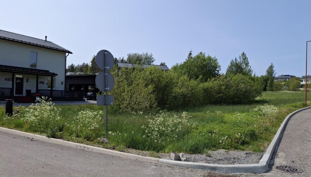 Valokuva rakentamattomasta tontista osoitteessa Uljaksentie 20. Tontilla kasvaa heinikkoa ja pensaita tai nuoria lehtipuita, vasemmassa laidassa näkyy naapurin omakotitalo.