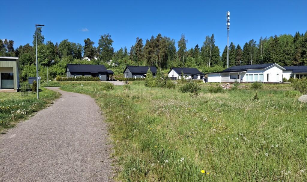 Ett foto av en äng där det finns fyra tomma tomter. Längs ängens vänstra kant går en parkgång, och längre bak syns enplans egnahemshus.