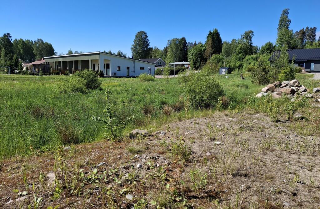 Ett foto. I förgrunden syns fyllnadsmassor på tomten vid Röjarevägen 10a, och bakom den ligger de tomma grannfastigheterna som täcks av vårgrön ängsvegetation. I bakgrunden finns enplans egnahemshus i grannskapet samt ett radhus.