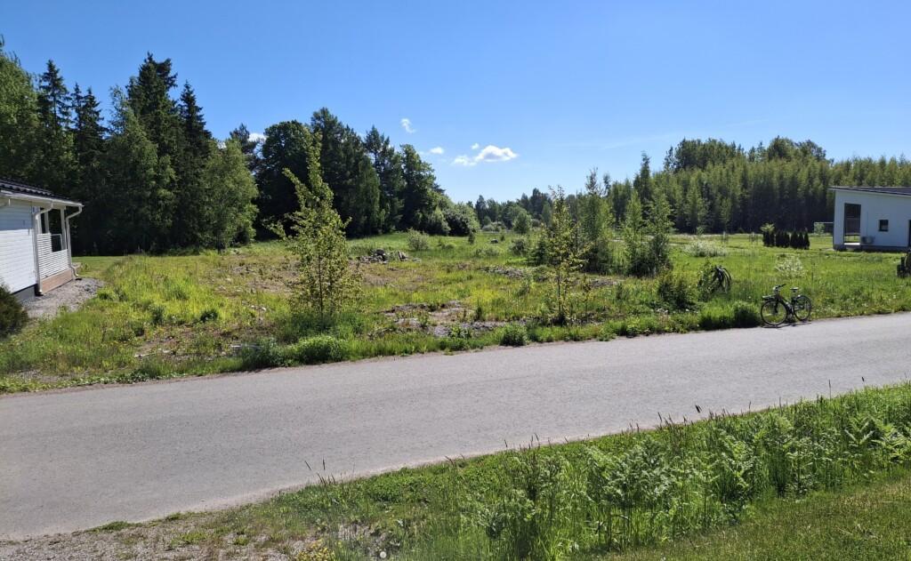 Ett foto. I förgrunden syns Röjarevägen. På andra sidan vägen ligger en äng där fyra tomma tomter finns. På båda sidor av bilden skymtar väggarna av grannhus, och bakom ängen står fullvuxna träd.
