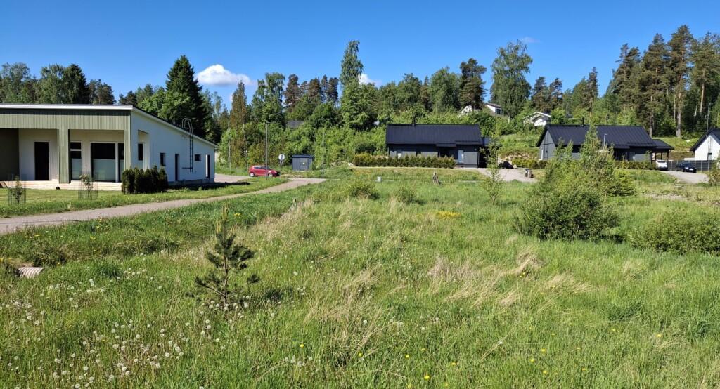 Ett foto av en äng där fyra tomma tomter ligger. På ängens vänstra sida löper en parkstig, och längre bak syns enplans egnahemshus samt gaveln av ett radhus.