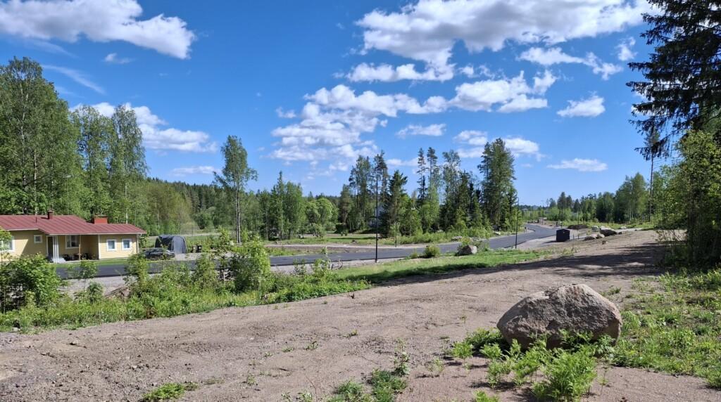 Valokuva. Etualalla rinteessä olevia tyhjiä tontteja, kuvan keskellä Skaftkärrintie, sen takana yksikerroksinen talo sekä lisää rakentamattomia tontteja ja alkukesän puustoa.