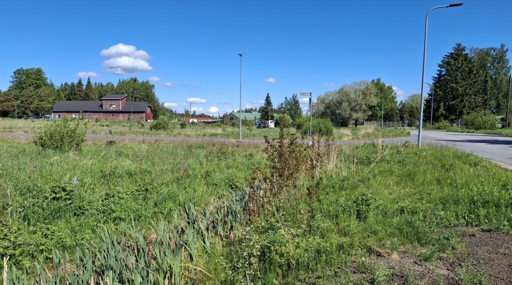 Valokuva niitystä, missä sijaitsee neljä tyhjää yritystonttia. Keskellä asfaltoitu Taitajankuja, niityn takana vanhoja rakennuksia ja oikeassa laidassa Pienteollisuustie.