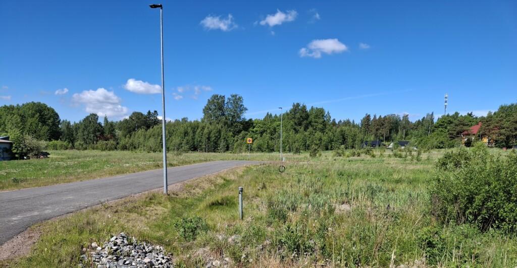 Valokuva niitystä, missä sijaitsee neljä tyhjää yritystonttia. Keskellä asfaltoitu Taitajankuja ja niityn takana täysikasvuista puustoa.