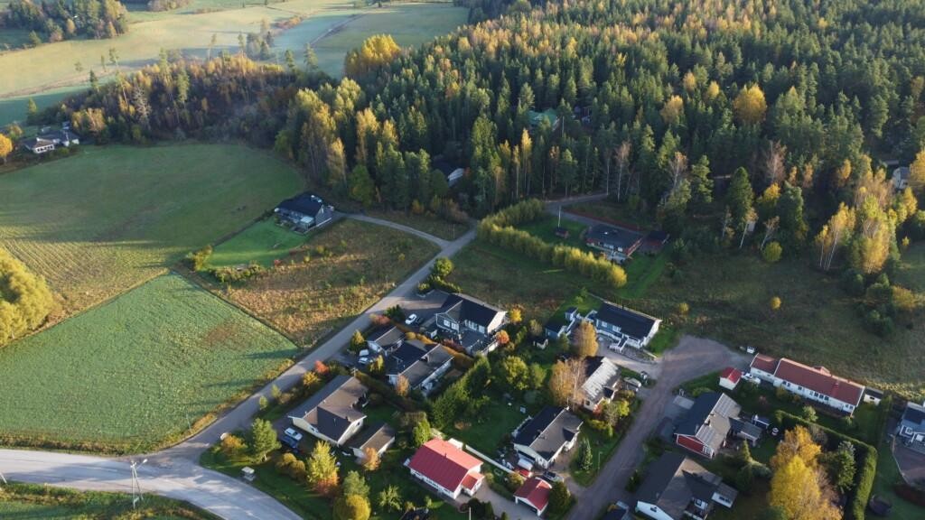 Flygfoto. Några egnahemshus med tillhörande gårdar, vägar, åkrar och skog. I mitten av bilden finns tre tomma tomter.