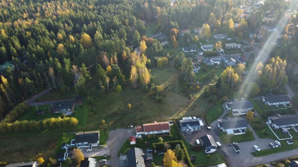 Flygfoto. På sluttningen, ungefär mitt i bilden, finns en tom tomt med träd som växer på ena sidan. Omgivningen består av egnahemshus och parkområde.