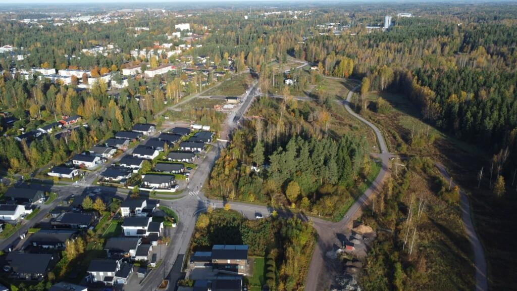 En omfattande flygfoto av kanten av ett växande bostadsområde i ett höstlandskap. På vänster sida av bilden syns småhus och gator, längre bort ett äldre bostadsområde. I mitten finns nya gator och obebyggda tomter. På höger sida och i bakgrunden finns ett stort skogsområde och enstaka byggnader.