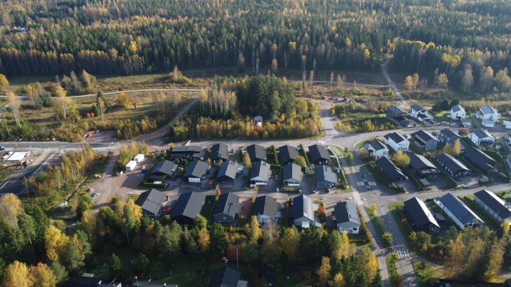 Flygfoto över det egnahems- och parhusdominerade bostadsområdet Vårdalen. I mitten av området finns egnahemshus, parhus, gator och gårdar. På vänster sida och längre bort finns tomma tomter. I bakgrunden breder ett stort höstfärgat skogsområde ut sig.