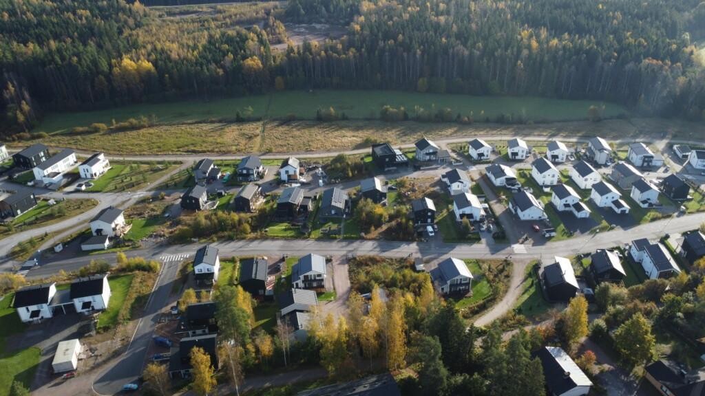 Flygfoto över bostadsområdet Vårdalen, som huvudsakligen består av egnahems- och parhus, med några enstaka obebyggda tomter här och där. Bakom bostadsområdet ligger en gång- och cykelväg, med en äng på ena sidan och skog bakom.