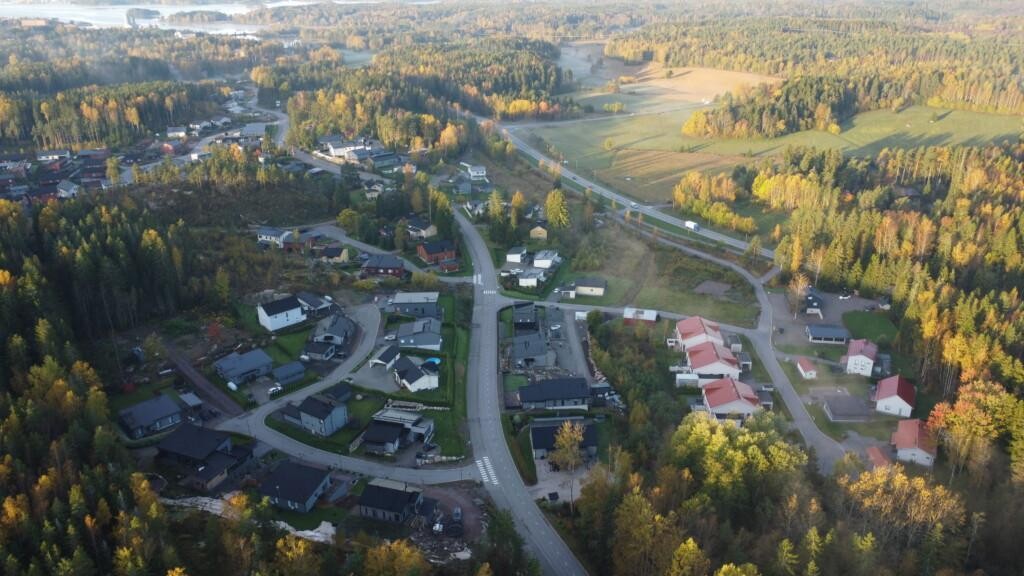 Flygfoto med egnahemshus och parhus samt gator och träd. I mitten av bilden finns några tomma tomter. Längre bort syns åkrar, skog och hav.