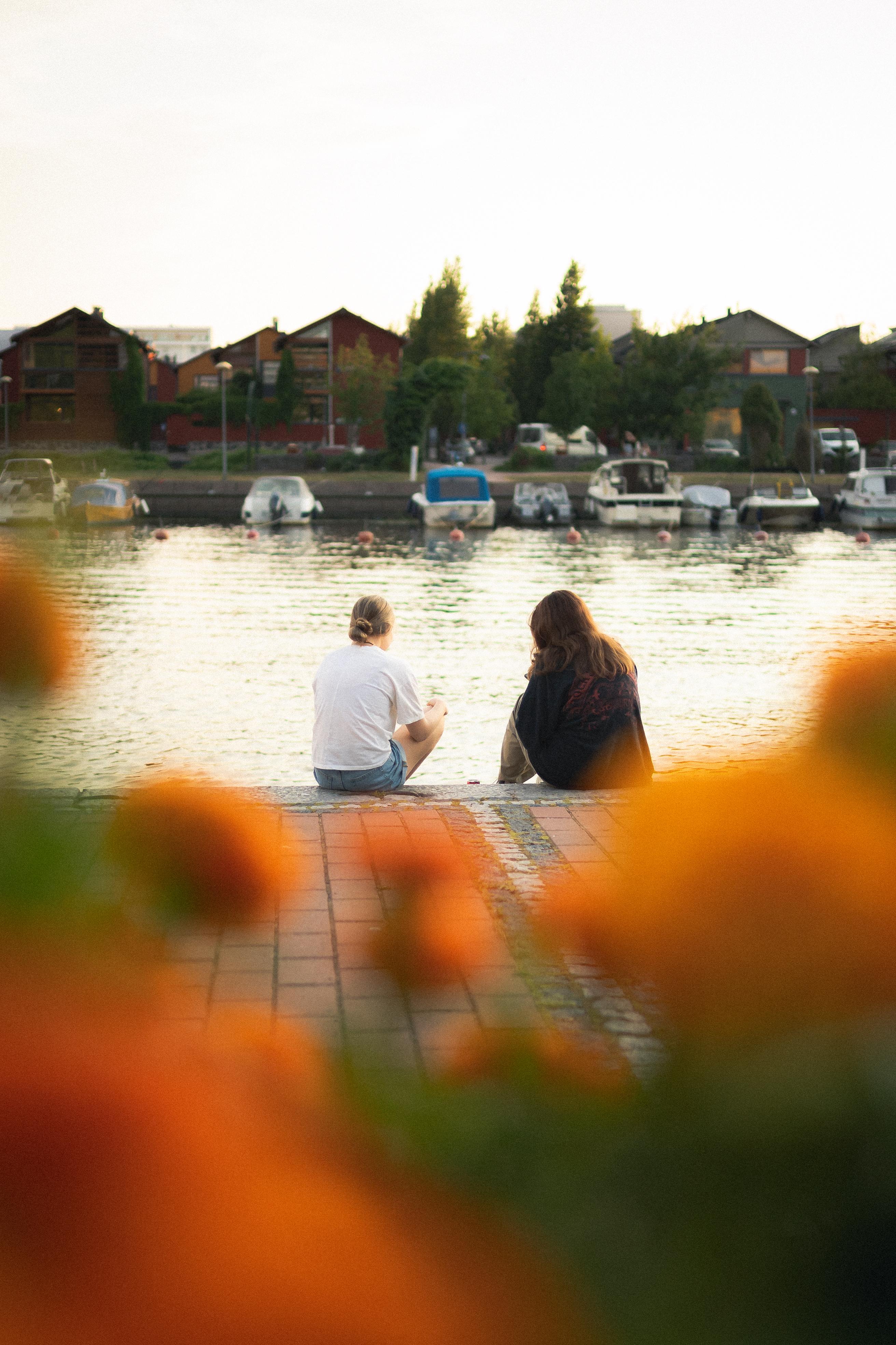Kukkia etualalla, taustalla Porvoon joen rannassa istuu kaksi nuorta, veneitä vastarannalla.