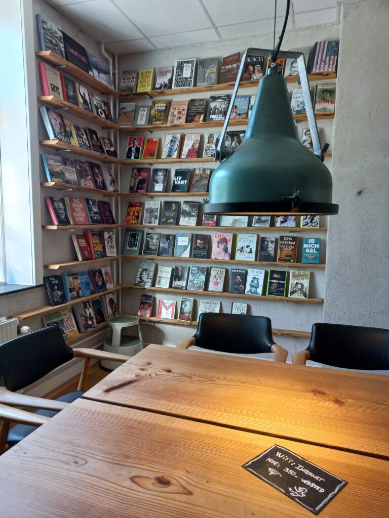Pöytä, tuoleja, valaisin ja kirjahylly kirjojen kannet esillä.