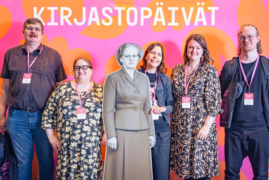 Viisi henkilöä ja yksi pahvifiguuri Kirjastopäivät-kyltin edessä.
