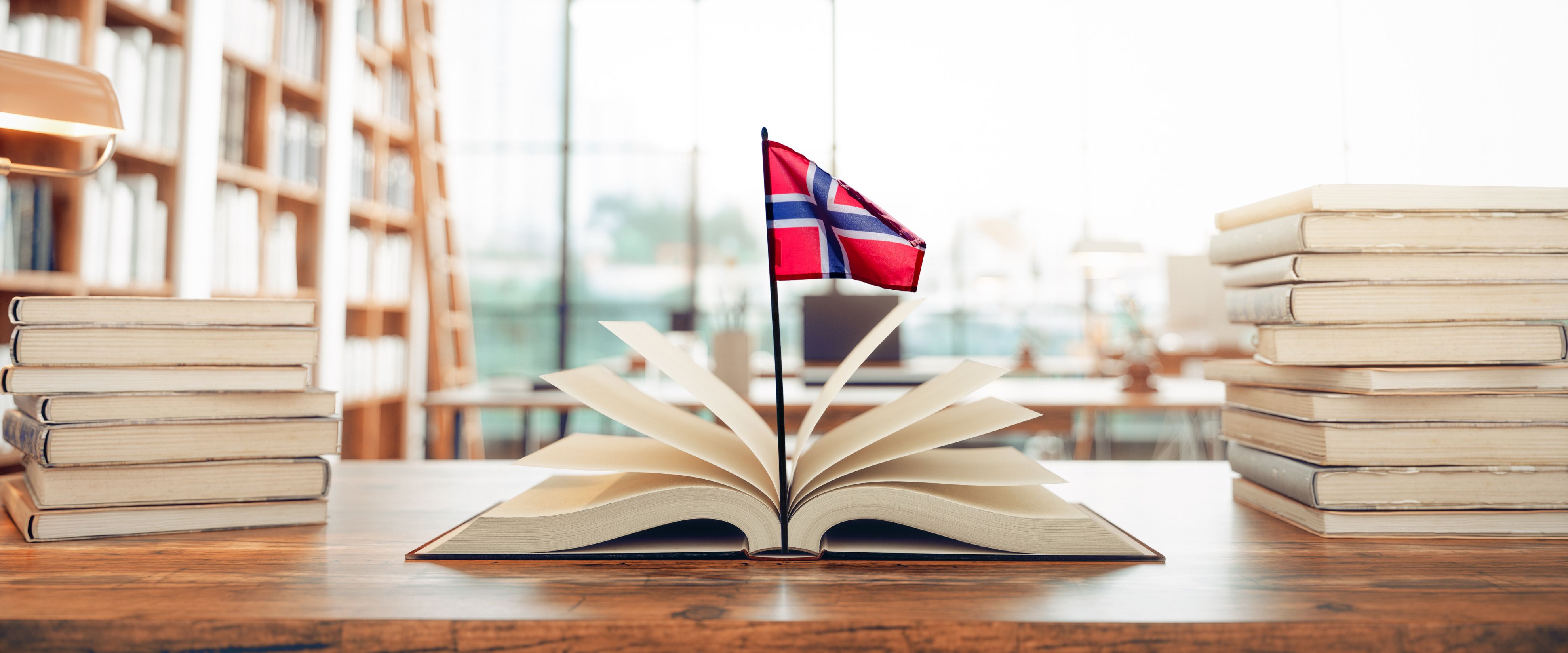 Kirjoja, kirjahyllyjä, avonainen kirja pöydällä ja sen päällä Norjan lippu.
