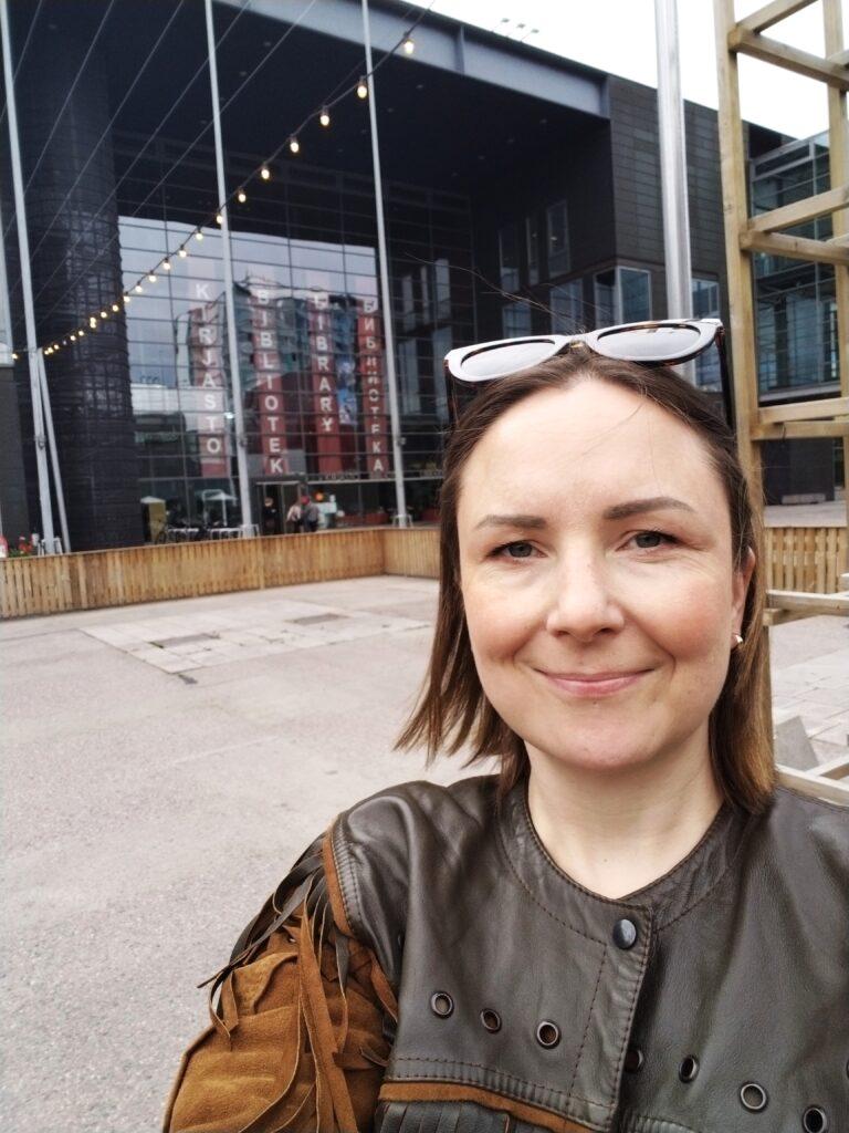 Hymyilevä henkilö kirjastorakennuksen edessä aurinkolasit päässään.