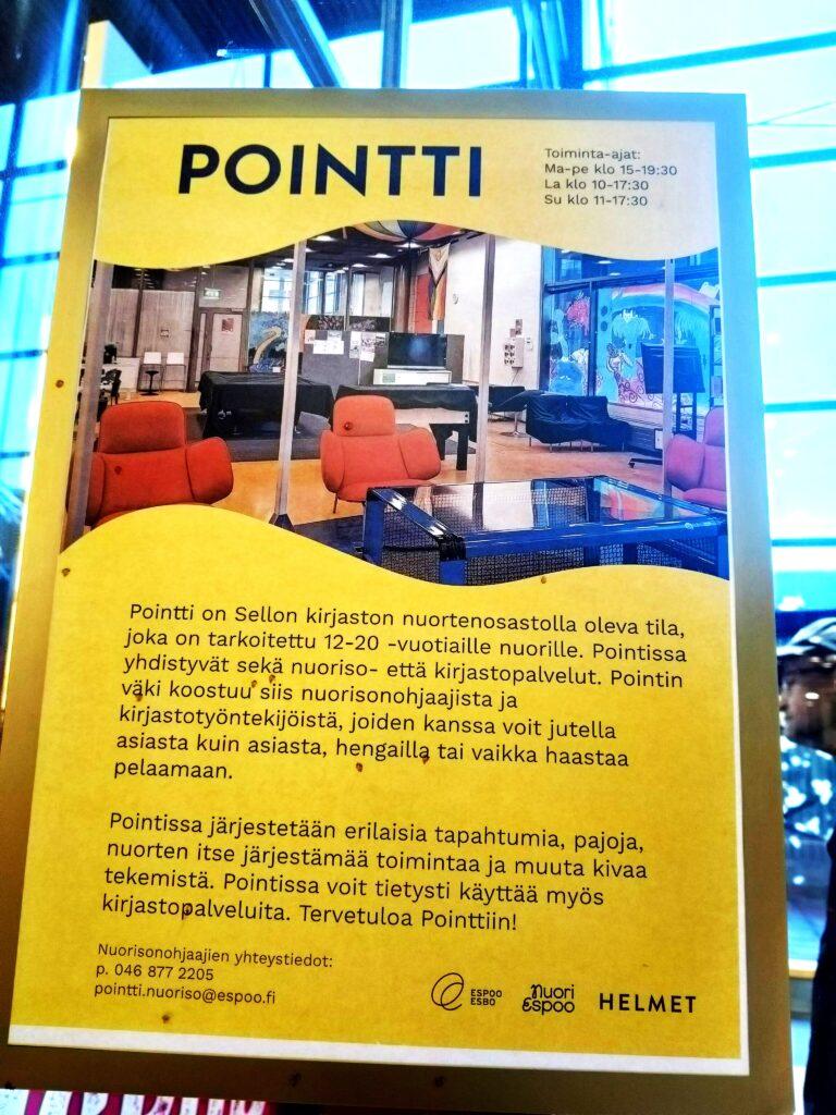 Keltainen Pointti-esittelykyltti Sellon kirjastossa. Kyltissä kerrotaan nuorille suunnatusta Pointti-tilasta, sen aukioloajoista ja toiminnasta. Kuvassa näkyy myös tilan sisustus punaisine nojatuoleineen.