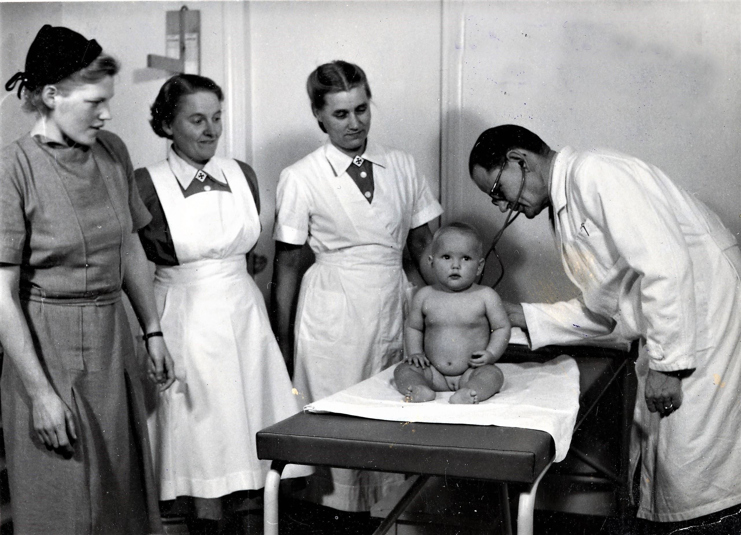 Mustavalkoinen kuva, jossa lääkäri tutkii vauvaa ja kolme naista seuraavat tilannetta.