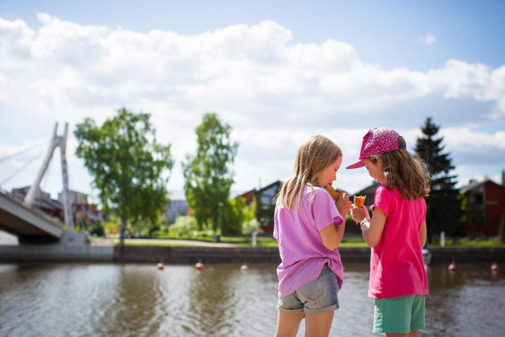 Valokuvassa kaksi tyttöä syö jäätelöä joen rannalla.