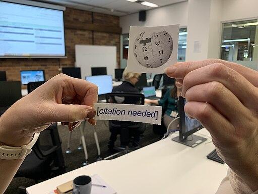 Henkilö pitää sormissaan paperilappuja joissa toisessa on Wikipedian logo ja toisessa [citation needed] teksti